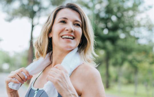 Smiling woman with blonde hair holding a towel around her neck while outdoors in a fitness setting.